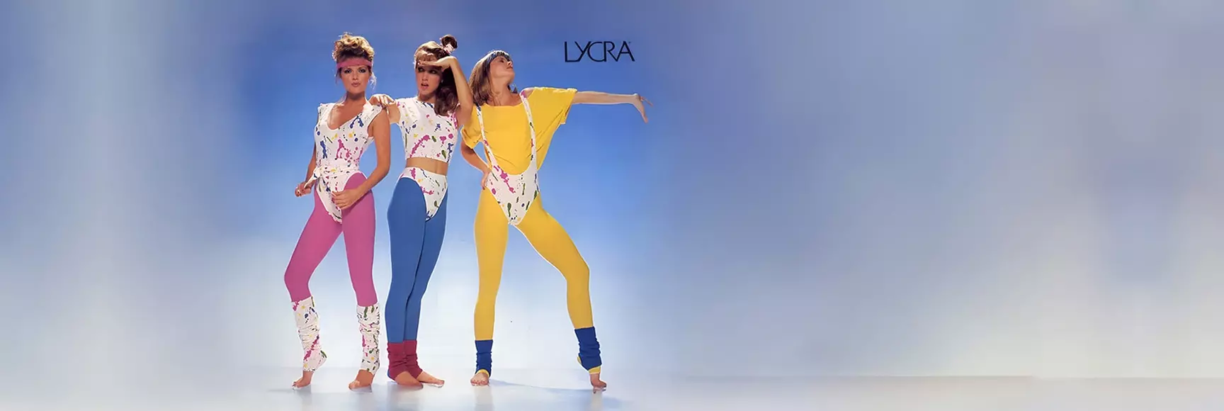 Three women in colorful ‘80s workout gear pose together on a blue background.